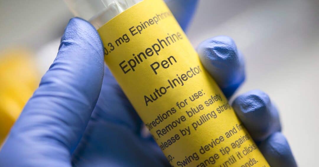 Close-up of a gloved hand holding an EpiPen autoinjector with a yellow label. The label includes the words "0.3 mg Epinephrine Pen" and "Auto-Injector" along with usage instructions.