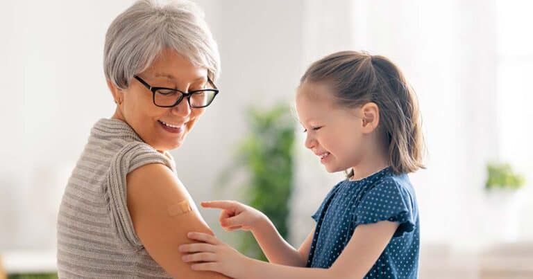 A smiling older woman with glasses and gray hair shows a bandage on her upper arm to a young girl with a ponytail, who is pointing at it. They are indoors, with a blurred green plant in the background.