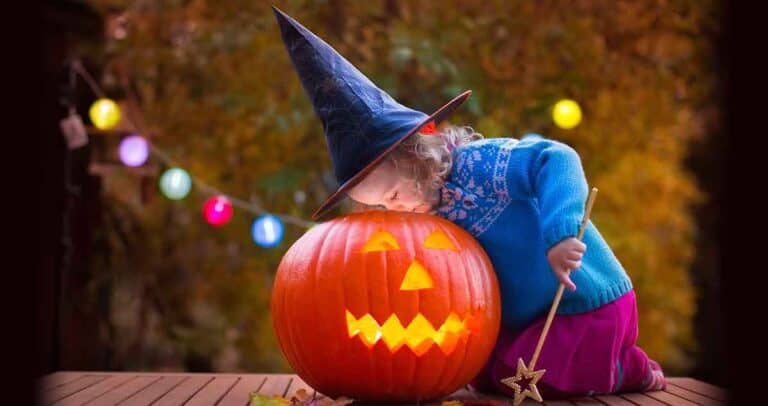 A child wearing a witch hat leans over a glowing jack-o'-lantern with a carved smile. They hold a wand and are dressed in a blue sweater and pink pants. Colorful lights hang in the blurred background, creating a festive Halloween atmosphere.