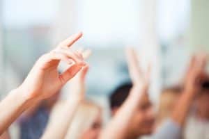 Close-up of several raised hands in a classroom or meeting setting, signifying people wanting to ask a question or participate actively. The background is blurred, highlighting the focus on the hands.