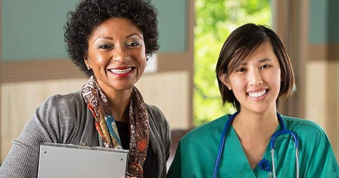 Two smiling women are indoors. One wears business attire and holds a folder; the other is in green medical scrubs with a stethoscope around her neck. They stand in a bright room with natural light streaming in.