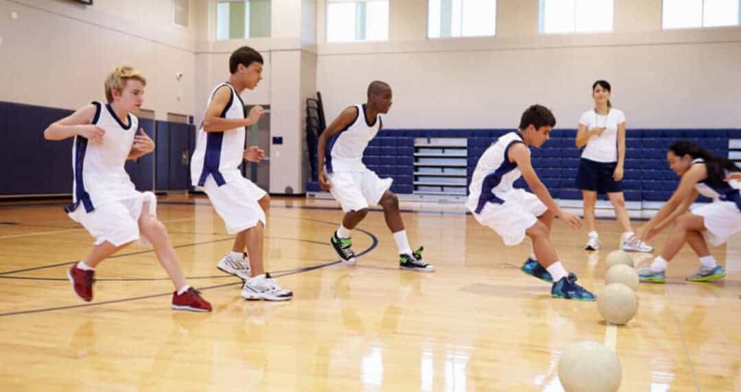 A group of four young athletes in blue and white uniforms engage in a basketball drill in a gymnasium, with a coach observing in the background. Two players are lunging toward basketballs on the floor.