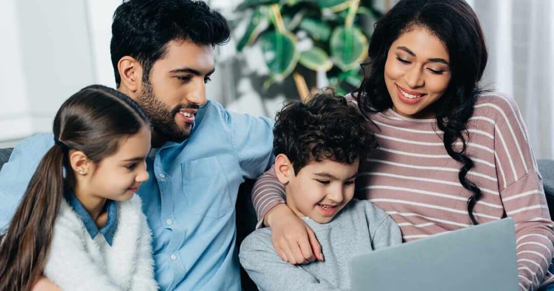 A family of four sits together on a couch, smiling and looking at a laptop. The man and woman sit on either side of two children, a girl and a boy. A large green plant is visible in the background.