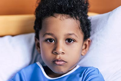 Ways to Give 1 A young child with curly hair wearing a blue shirt looks directly at the camera while lying on a bed with white pillows and a wooden headboard. The expression is calm and thoughtful.