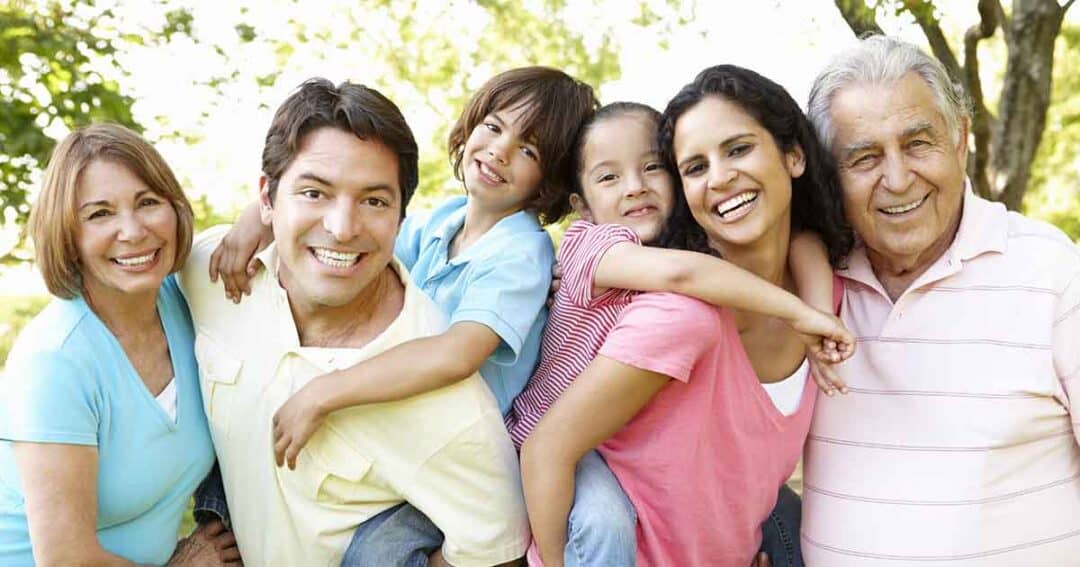 A happy family of six stands outdoors, smiling. Two children are being playfully held by a man and woman, surrounded by an older couple. They wear casual summer clothes, and trees are visible in the background.