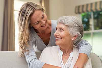 Ways to Give 2 A young woman with long blonde hair smiles while embracing an older woman with short gray hair. They are indoors, sitting on a couch in a cozy room.