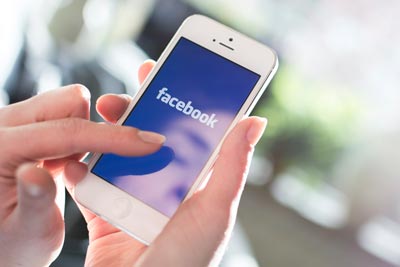 Ways to Give 3 A person holding a smartphone displaying the Facebook app on the screen, with their thumb poised to tap. The background is softly blurred.