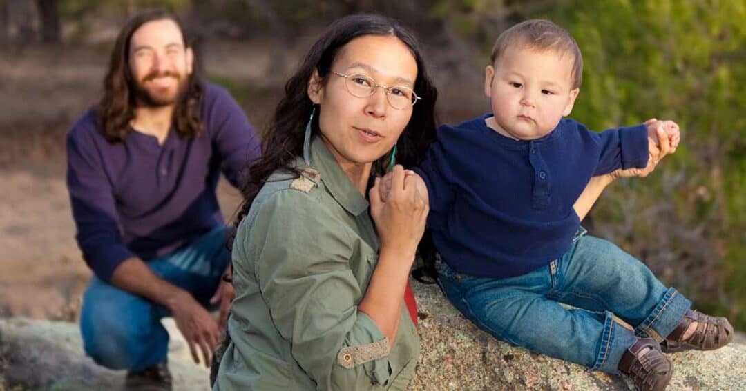A mother holding a baby boy sits on a rock outdoors, looking at the camera. The baby is wearing a blue shirt and jeans. A man with long hair and a beard kneels in the background, smiling. Trees are visible in the background.