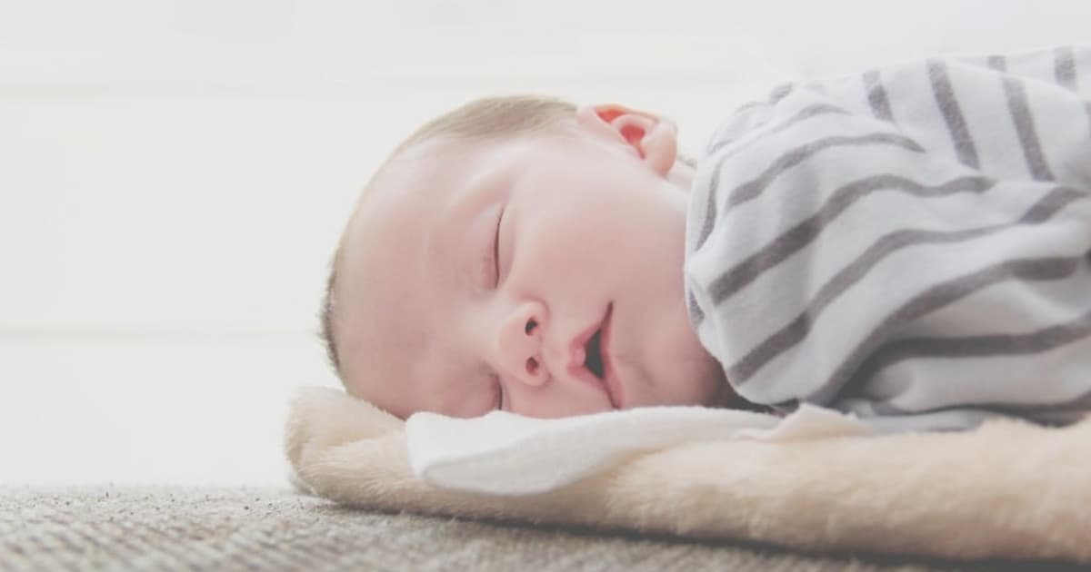 A sleeping baby lies on a soft surface, wrapped in a striped blanket. The baby has a peaceful expression, and the setting is softly lit, creating a cozy and serene atmosphere.