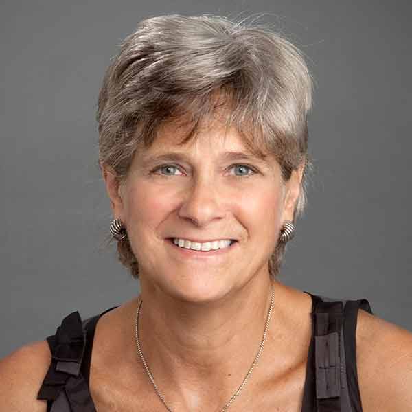 A person with short gray hair smiles at the camera. They are wearing a sleeveless black top, silver hoop earrings, and a silver necklace. The background is a plain gray.