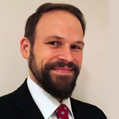Man with a beard smiles at the camera, wearing a dark suit, white shirt, and a red tie with blue dots, standing against a plain background.