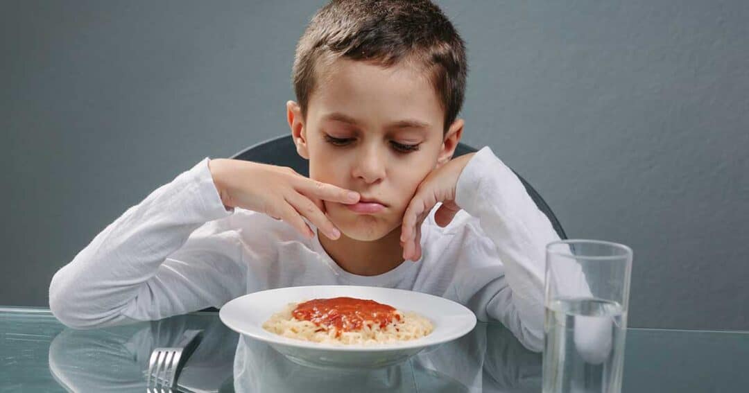 A young boy in a white shirt stares anxiously at a plate of spaghetti with tomato sauce, worried about possible food allergies. He sits at a glass table with a glass of water nearby, set against a plain gray background.
