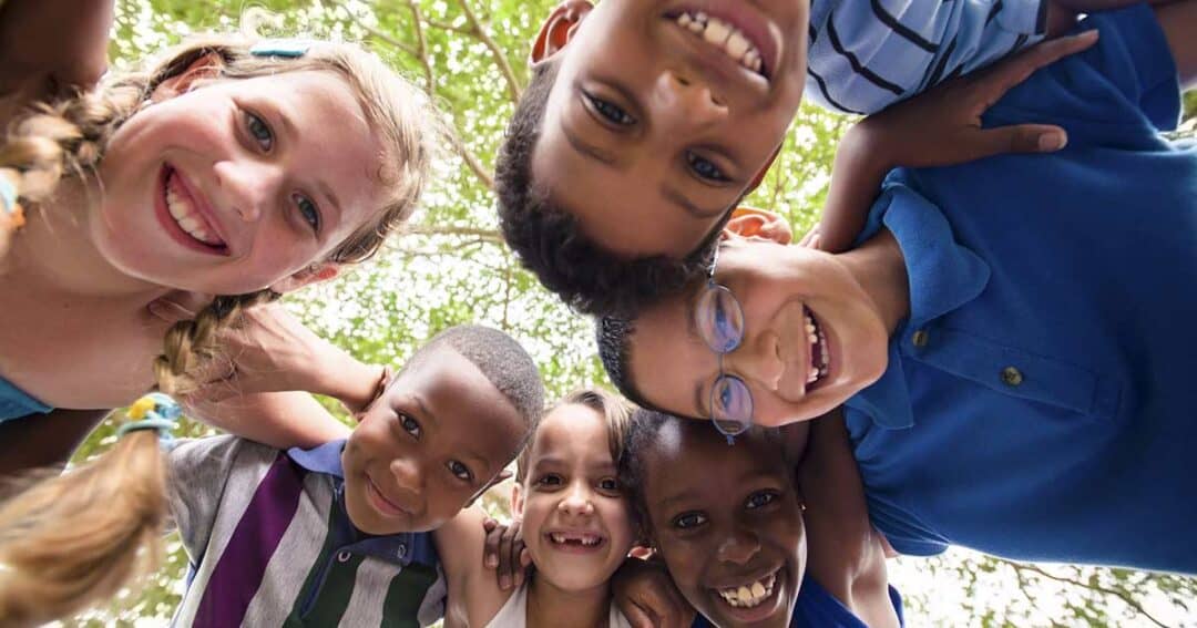 A group of six diverse, smiling children form a circle and look down at the camera. They are outdoors, with green trees in the background, creating a joyful and playful atmosphere. The children are leaning in close, bonding happily.