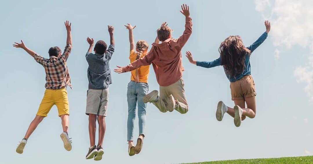 Five people are joyfully jumping in the air on a grassy hill against a clear blue sky. They are facing away from the camera, dressed in casual summer clothes, and their arms are raised high. The scene conveys a sense of freedom and happiness.