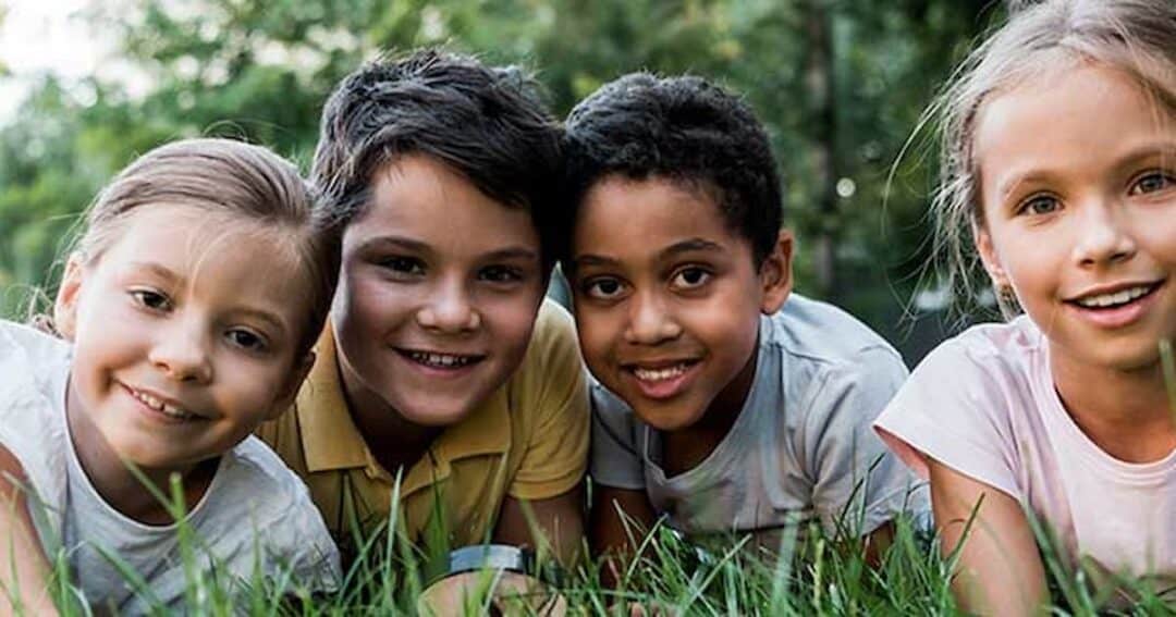 Grass Allergy 6 Four children are lying on the grass, smiling at the camera. The scene is set outdoors with greenery in the background. They appear happy and relaxed, enjoying a moment together in nature.
