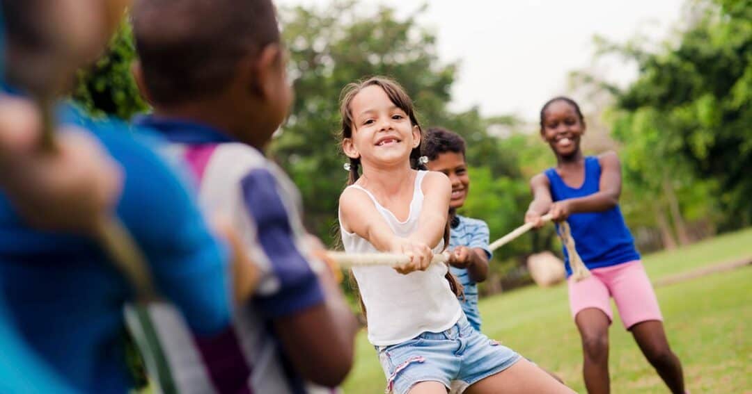 Children smiling and playing tug-of-war on a grassy field. The focus is on a young girl in a white tank top, pulling the rope with determination. Other children in colorful clothes are also pulling, with greenery in the background.