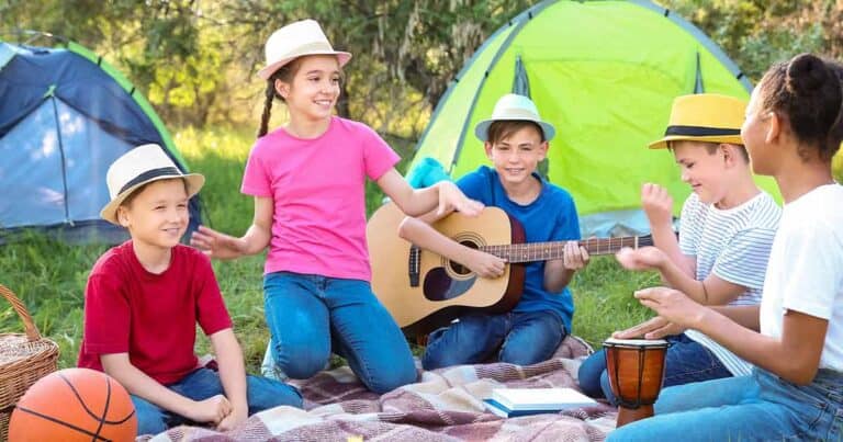 Five kids sit on a blanket outdoors near tents, wearing hats. One plays guitar, another plays a drum, and they all smile and interact during their camping trip, where having epinephrine at summer camp ensures everyone stays safe while having fun.