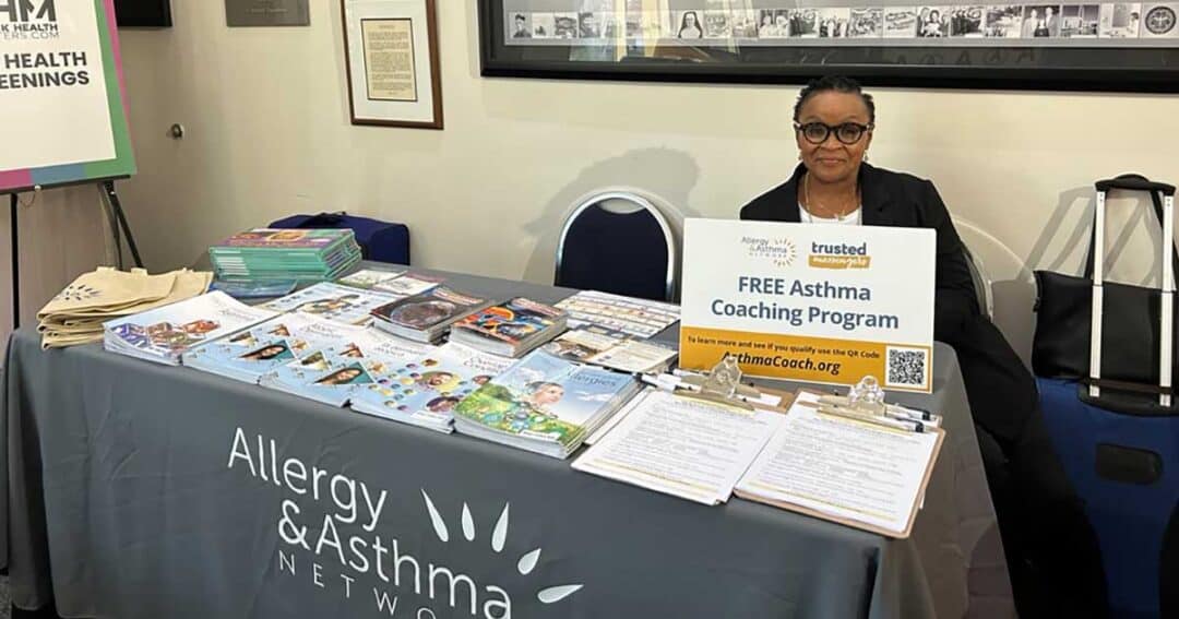 Laonis Quinn RN, MSN, AE-C sits at an information booth with a sign that reads "FREE Asthma Coaching Program." The table is covered with brochures, magazines, and clipboards. A banner behind the table proudly displays "Allergy & Asthma Network.