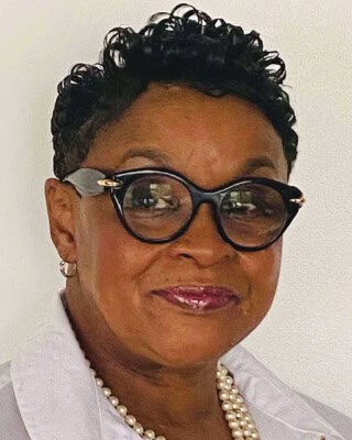 A person wearing black-framed glasses, a pearl necklace, and a white collar smiles slightly at the camera, set against a plain background.