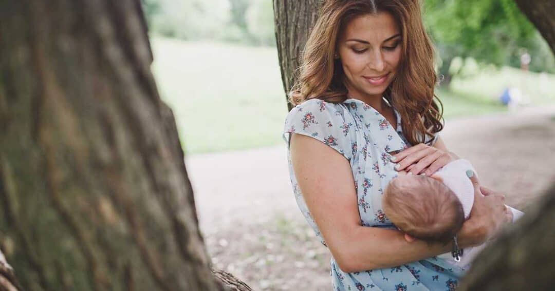 A woman with long brown hair wearing a floral blouse is smiling down at a baby she is holding. They are outdoors, surrounded by trees and greenery.