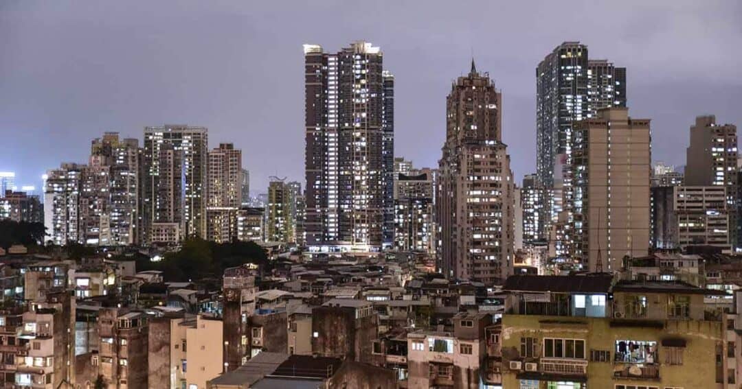 A nighttime cityscape featuring a mix of modern skyscrapers and older buildings. The skyscrapers are brightly lit, contrasting with the dimly lit shanty town in the foreground. The sky is overcast, intensifying the wealth disparity in the area.