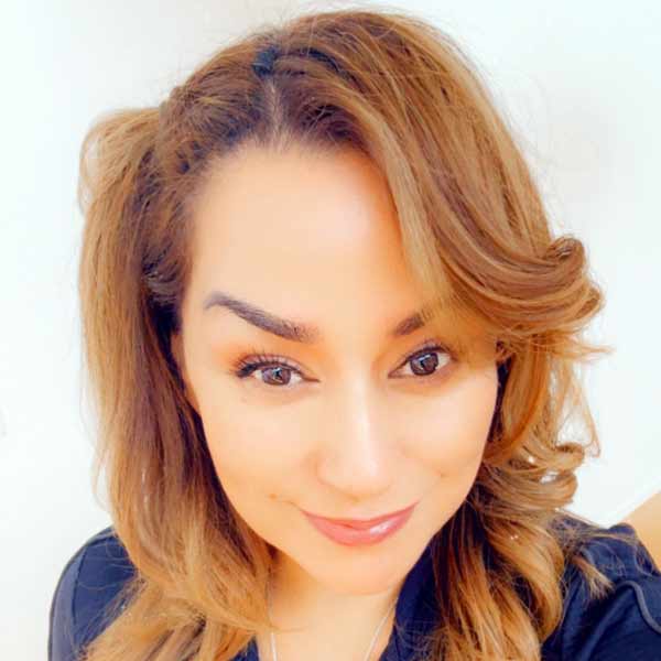 A woman with wavy, light brown hair and subtle makeup smiles softly at the camera. She is wearing a dark top and is positioned against a plain light background.