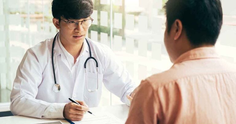Your Trusted Resource 19 A doctor with glasses and a stethoscope around his neck sits at a desk, holding a pen and talking to a patient who is seated opposite him. The setting is a bright office with frosted windows in the background.