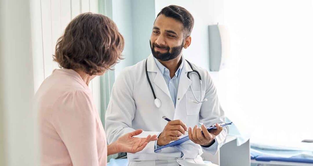 A doctor in a white coat and stethoscope smiles and takes notes on a clipboard while talking to a patient with short brown hair, who gestures expressively. They are sitting in a brightly lit medical office.