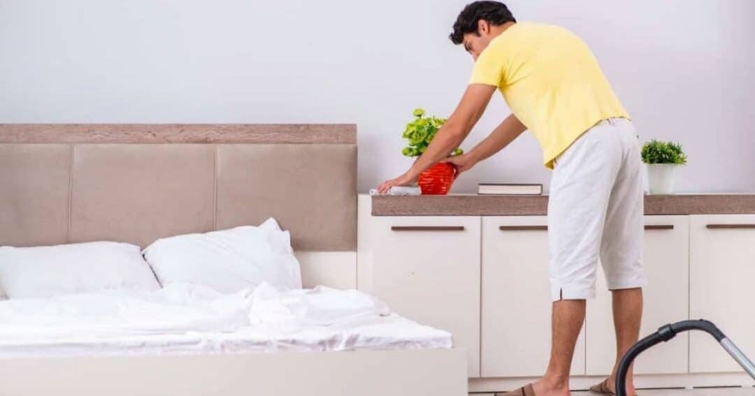 A man in a yellow shirt and white pants tidies a dresser beside an unmade bed. A red vase with a plant and a book are on the dresser. A vacuum cleaner is on the floor nearby. The room has a minimalist design.