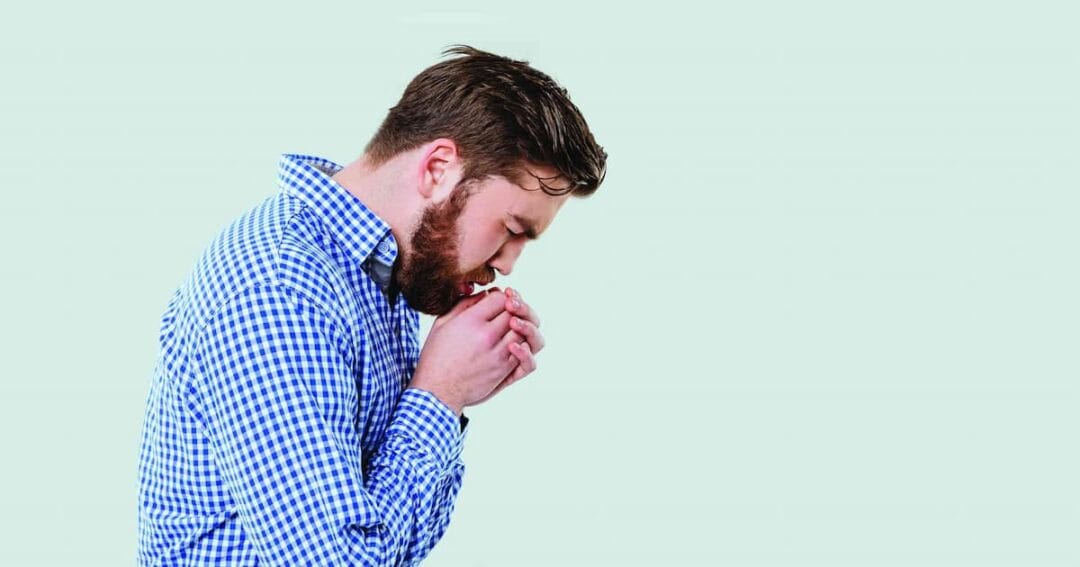 A man in a blue checkered shirt is shown in profile, appearing to cough or sneeze into his hands. The background is a plain light green.