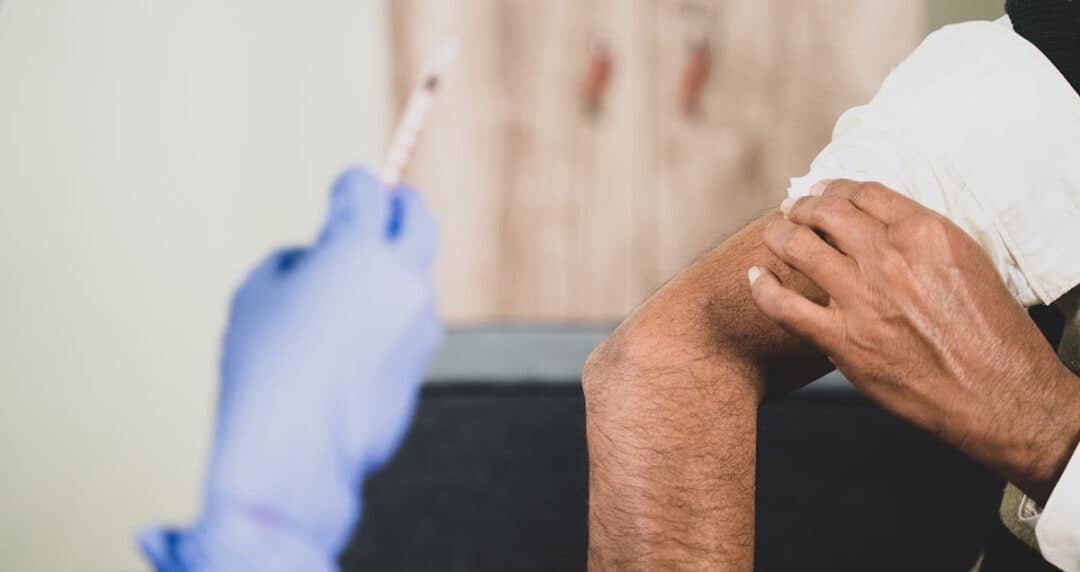A person receives an allergy shot in their upper arm. A gloved hand holds a syringe on the left, while the person rolls up their sleeve on the right. The background is blurred and neutral-toned.