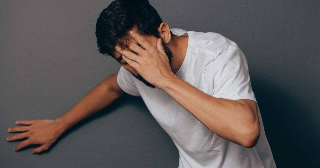 A man in a white shirt leans against a gray wall, covering his face with one hand, as if warding off the onset of anaphylaxis. His expression reveals distress or headache, underscoring the urgency of his situation.