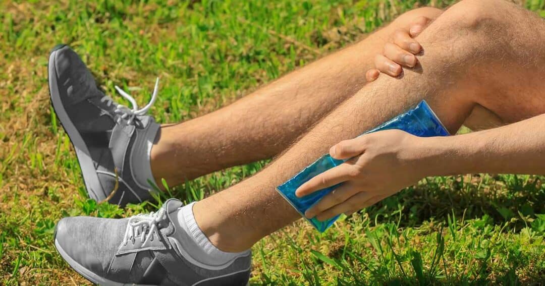 A person wearing gray sneakers applies a blue ice pack to their left shin while sitting on grass. They are holding their leg with the other hand, suggesting relief or treatment for pain or injury.