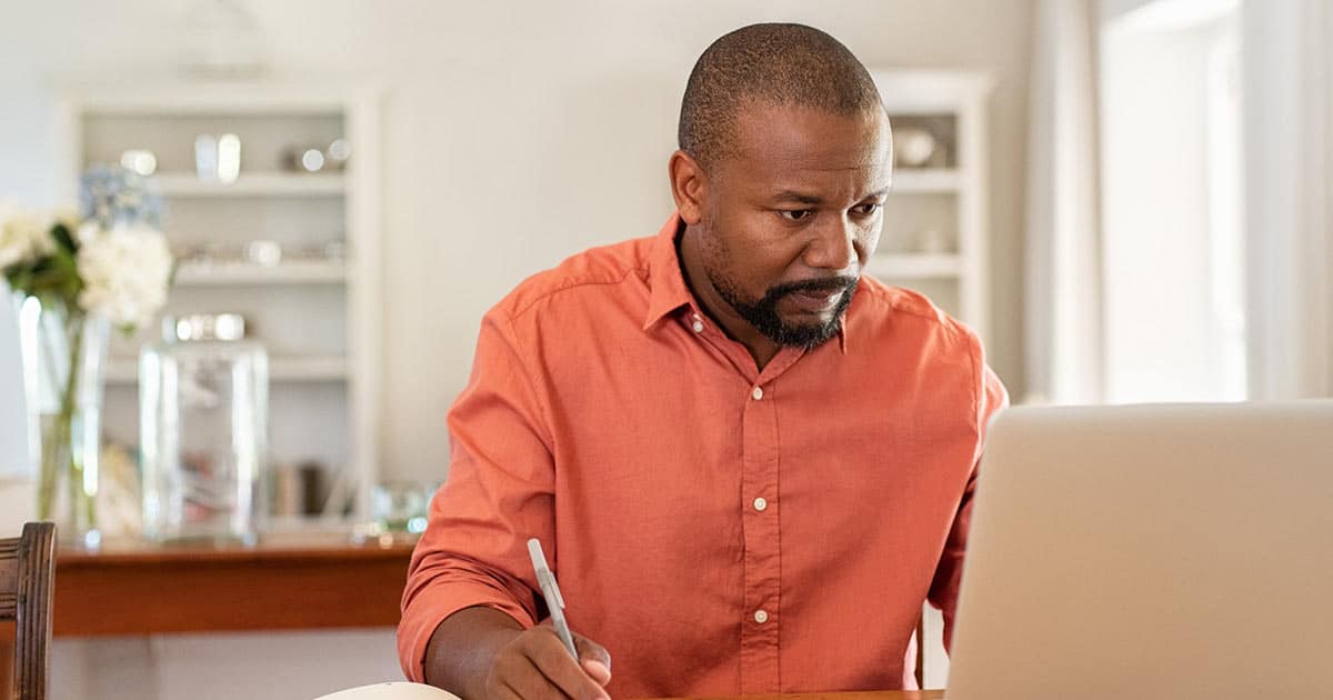 Resources for Care and Support 6 A man in an orange shirt sits at a table, focused on his laptop. He holds a pen in one hand. The background shows a bright room with shelves and a vase of flowers.