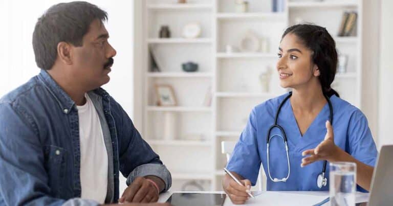A healthcare professional in blue scrubs, with a stethoscope around her neck, consults with a man in a denim jacket. They are sitting at a table with a notepad, tablet, and glass of water in a bright, airy room.