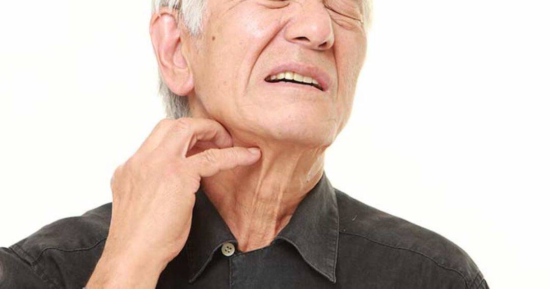 An older man wearing a dark shirt touches his neck and winces, showing discomfort or pain against a plain white background.