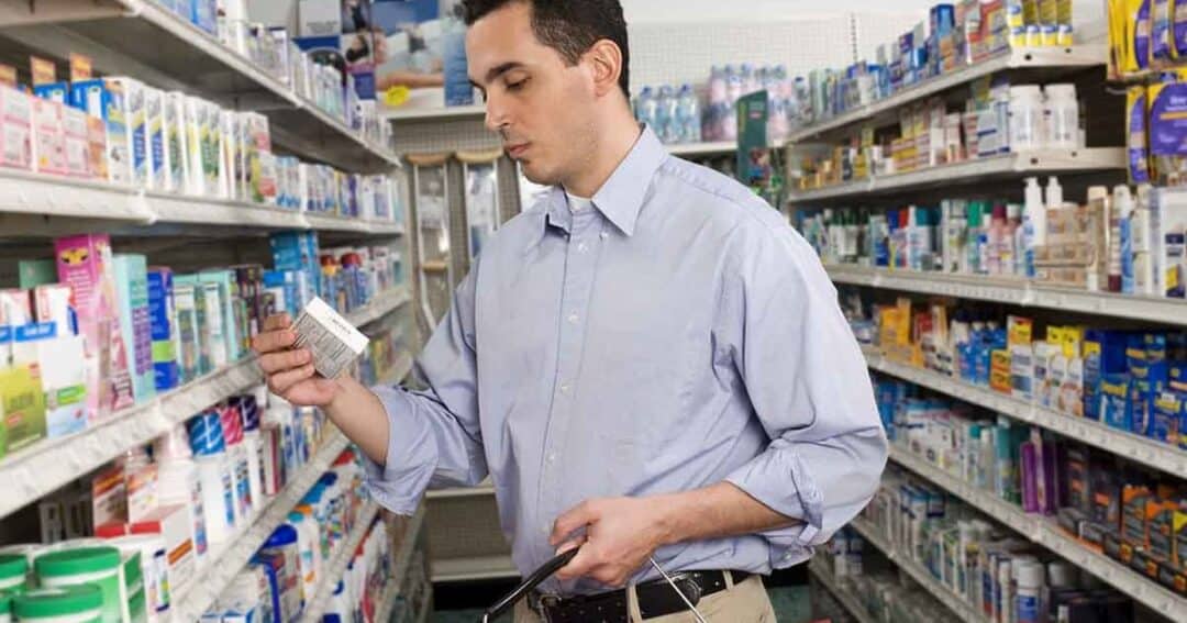 What if I Can’t Afford My Allergy Medications? 1 A man in a blue shirt is standing in a pharmacy aisle, holding a product and reading its label. Shelves stocked with various health products surround him. He is carrying a shopping basket.