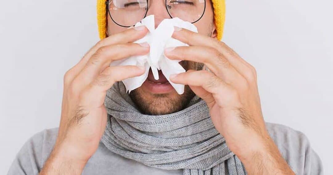 A person wearing glasses, a yellow beanie, and a gray scarf is holding a tissue to their nose, appearing to sneeze or blow their nose. The background is plain and light-colored.