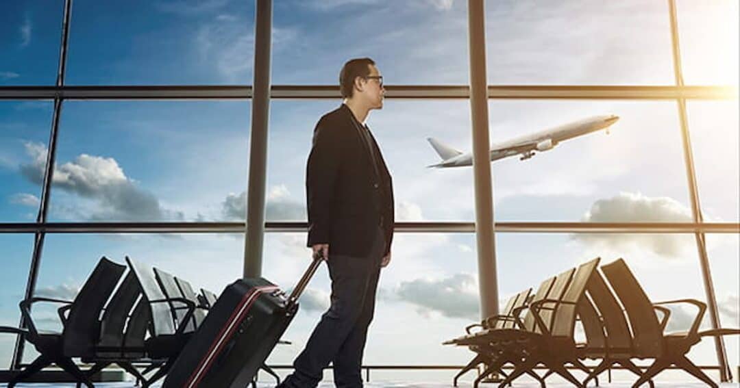 A man in a suit walks through an airport terminal, pulling a suitcase. Rows of empty seats are visible, and a plane is seen taking off through large windows framing a bright sky.