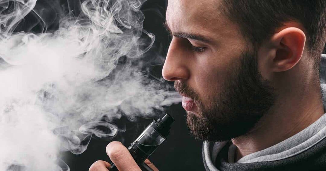 A man with a beard exhales vapor while holding a vape device near his mouth. Thick clouds of vapor surround his face against a dark background. He is wearing a hoodie and appears focused on vaping.