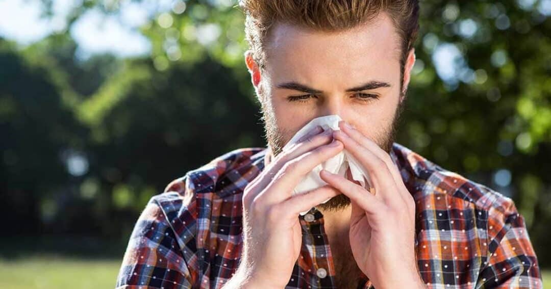 Grass Allergy 12 A man with a beard wearing a plaid shirt stands outdoors, holding a tissue to his nose as if sneezing or blowing his nose. Behind him are blurred trees and greenery, suggesting he is in a park or natural setting.