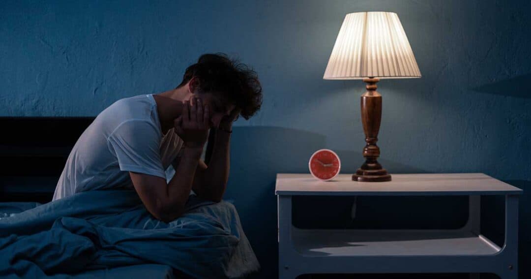 Man with nocturnal asthma A person sits on a bed in a dimly lit room, head in hands, appearing thoughtful or stressed. A lamp glows on a bedside table next to an alarm clock. Battling nocturnal asthma under the calming yet somber blue walls adds to the night's quiet struggle for breath.