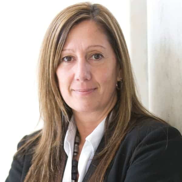 A woman with long brown hair, wearing a black blazer and white shirt, stands against a light-colored wall. She is looking at the camera with a slight smile.