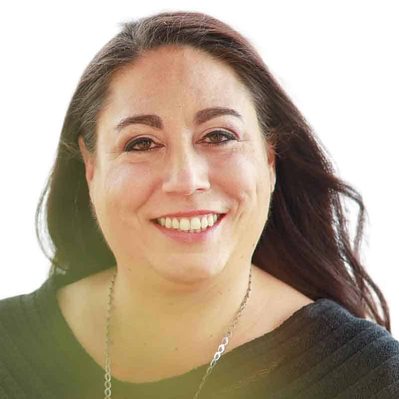 A person with long dark hair smiling, wearing a black top and a necklace, against a plain white background.