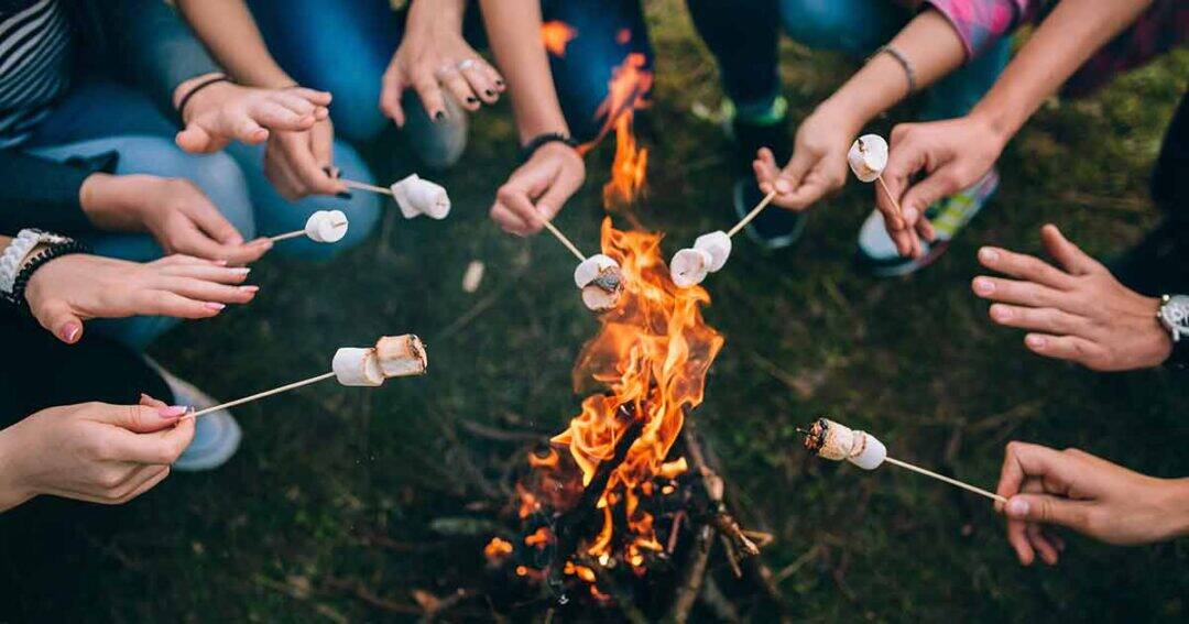 A group of people sitting around a campfire at summer camp, roasting marshmallows on sticks over the flames. Only their hands and sticks are visible, with epinephrine kits nearby as the fire burns brightly in the center.