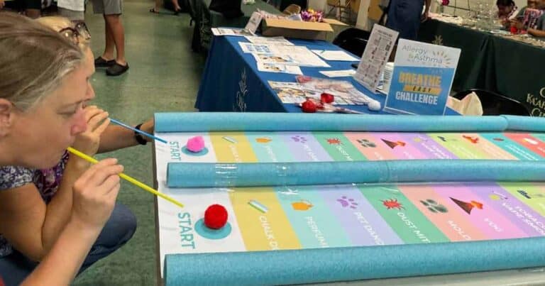 A woman blows a red pom-pom along a colorful game board using a straw at a table display. The board has labeled sections, foam barriers, and a "Breathe Easy Challenge" sign relating to asthma and allergies.