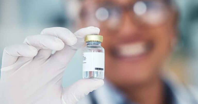 A person wearing glasses and white gloves smiles while holding up a small vial with a white label and a gold cap. The background is blurred, drawing focus to the vial and the person's enthusiastic expression.