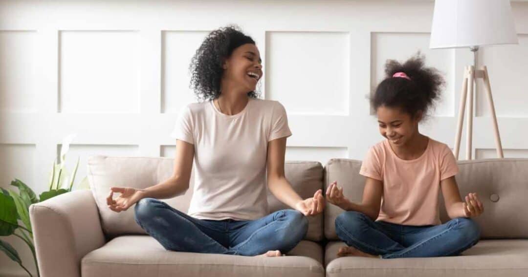 A woman and a young girl sit cross-legged on a sofa, smiling as they meditate together. They both wear casual shirts and jeans. The room is softly lit with a white wainscoted wall and a floor lamp in the background.