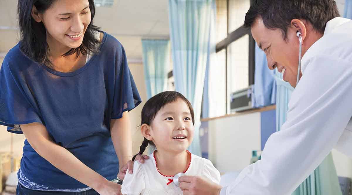 A doctor examines a smiling child with a stethoscope. The child, wearing a red necklace and a white shirt, is sitting next to a woman who is gently holding them. The setting appears to be a hospital or clinic room.