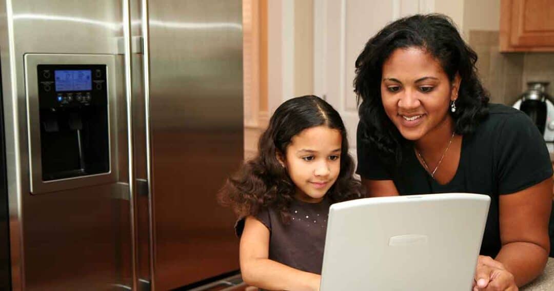 A woman and a young girl are smiling while looking at a laptop in a kitchen. They stand next to a stainless steel refrigerator. The woman is helping the girl, suggesting a teaching or learning activity.